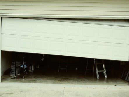 07-small Obraz na stronie internetowej przedstawia jasny garaż z częściowo otwartymi i źle ustawionymi drzwiami, które wyglądają na poza torem. Przez otwór widać zagracone wnętrze wypełnione różnymi przedmiotami gospodarstwa domowego i narzędziami.