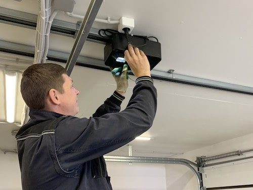 11 Na stronie internetowej poświęconej naprawie bram, rolet i barier w Warszawie, na zdjęciu widać technika w ciemnej kurtce pracującego nad instalacją lub regulacją otwieracza drzwi garażowych zamontowanego na suficie garażu. Scena obejmuje widoczne szczegóły, takie jak przewód zasilający i częściowo otwarte drzwi garażowe w tle.