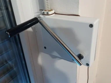 12519b464aa0a56c9ee0e90cdc8d Obraz na stronie internetowej przedstawia białą prostokątną skrzynkę sterowniczą zainstalowaną na ścianie, obok okna. Do niej przymocowana jest przedłużona ręczna korba z czarnym uchwytem, przeznaczona do obsługi bram, rolet lub barier w Warszawie.