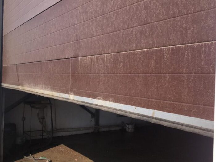 1620064841797-1024x576 Obraz na stronie internetowej przedstawia dużą, przemysłową bramę garażową w Warszawie, częściowo podniesioną, aby odsłonić nieskazitelną betonową podłogę w środku. Zewnętrzna część bramy ma brązowy odcień ze śladami zużycia i jest zaakcentowana pomarańczowymi i białymi obramowaniami. Po lewej stronie ramy widać część pojazdu budowlanego.