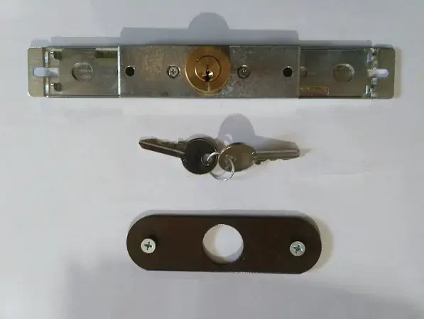 2095783775_w600_h600_2095783775 Obraz na naszej stronie internetowej, poświęconej naprawom bram, rolet i barier w Warszawie, przedstawia szczegółowy widok metalowego mechanizmu zamka drzwi. Mechanizm ten obejmuje cylindryczny zamek wraz z dwoma towarzyszącymi mu kluczami, starannie przymocowanymi do breloka. Pod zamkiem znajduje się ciemna prostokątna płytka z elegancko zaprojektowanym okrągłym wycięciem. Wszystkie elementy są starannie ułożone na gładkiej białej powierzchni, aby zapewnić wyraźną widoczność.