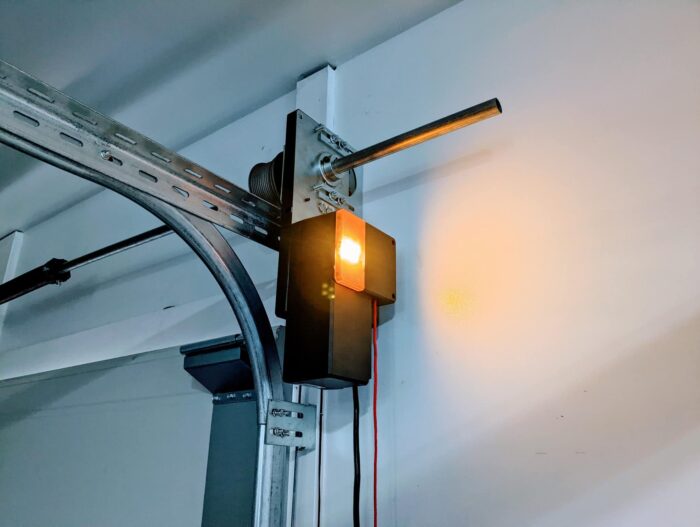 325593434_574780547902631_6769590877031841360_n Obraz na stronie internetowej naprawy bram w Warszawie przedstawia oświetloną oprawę świetlną przymocowaną w pobliżu elementów mechanicznych szyny bramy garażowej. Ciepłe światło, które emituje, delikatnie oświetla białą ścianę, podkreślając szynę i jej metalowe okucia, wyraźnie widoczne w scenie.