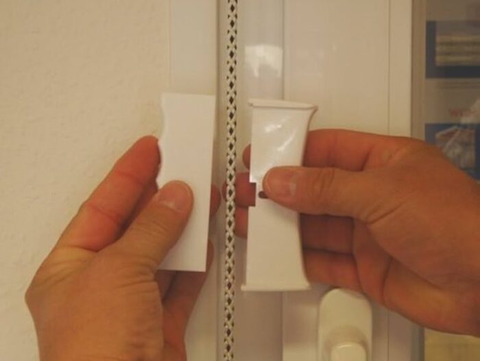 41NHZLKOO0L._AC_UF894,1000_QL80_ Obraz na stronie internetowej przedstawia zbliżenie dwóch rąk, które skrupulatnie pracują nad dopasowaniem kawałków białej osłony zawiasu do metalowego zawiasu wahadłowego, który jest przymocowany do ramy drzwi lub okna. Po prawej stronie ramy wyraźnie widoczna jest klamka, podkreślająca szczegółowe rzemiosło związane z naprawą bram, rolet i barier w Warszawie.