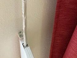 61WmUrH9fdL._UX250_ Obraz na stronie internetowej przedstawia teksturowaną beżową ścianę z białą karniszem bezpiecznie zamontowanym. Do karnisza przymocowany jest pojedynczy haczyk, a czerwona zasłona częściowo opada, widoczna po prawej stronie ramy.