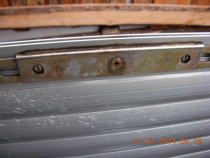 9e05494s-960 Przedstawiono szczegółowy widok metalowego mechanizmu zamka na falistych drzwiach przesuwnych. Zamek ma dwie śruby po każdej stronie i wykazuje niewielkie oznaki zużycia. Zarówno drzwi, jak i otaczający je obszar wykazują zauważalne ślady zużycia, wskazujące na lata użytkowania. Pieczątka z datą u dołu wskazuje „27.03.2014 09:36”, sugerując, kiedy zrobiono to zdjęcie lub kiedy ostatnio odnotowano konserwację.