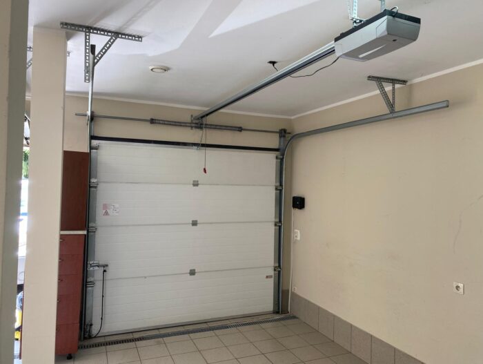 WhatsApp Image 2024-07-19 at 14.12.48 Obraz na stronie internetowej przedstawia nieskazitelny, niezamieszkany garaż z zamkniętymi białymi drzwiami garażowymi. Sufit jest wyposażony w automatyczny system otwierania drzwi dla wygody. Podłoga jest elegancko wyłożona kafelkami, a ściany mają uspokajający beżowy odcień. Po jednej stronie znajduje się szafka do przechowywania, a sufit jest wyposażony w kompaktową oprawę oświetleniową, aby oświetlić przestrzeń.