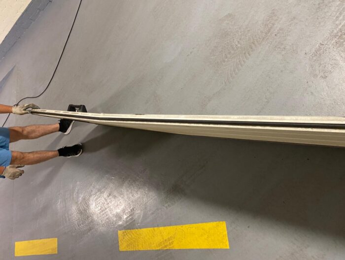 WhatsApp Image 2024-07-28 at 14.29.37 Obraz na stronie internetowej przedstawia pracownika ubranego w niebieskie spodenki, czarne trampki i rękawice robocze, który sprawnie obsługuje długą, elastyczną blachę na szarej podłodze w pomieszczeniu. Na podłodze widać żółte oznaczenia, gdy osoba ta rozwija blachę. Ta scena jest częścią ich zadania związanego z naprawą bram, rolet i barier w Warszawie.