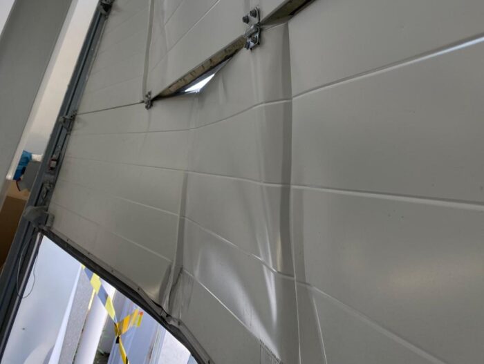 WhatsApp Image 2024-07-28 at 14.53.50 Obraz na stronie internetowej poświęconej naprawom w Warszawie przedstawia białą bramę garażową, która jest częściowo otwarta, odsłaniając widoczne wgniecenia i zagięcia wzdłuż pionowych paneli. W tle widać żółtą taśmę ostrzegawczą, sygnalizującą obszar wymagający uwagi lub naprawy.