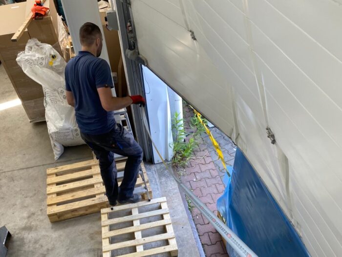 WhatsApp Image 2024-07-28 at 14.54.20 W otoczeniu przemysłowym przedstawionym na warszawskiej stronie internetowej poświęconej naprawom bram, rolet i barier, technik w niebieskiej koszuli i czerwonych rękawiczkach umiejętnie obsługuje częściowo otwarte drzwi garażowe. Są one umieszczone na drewnianych paletach obok dużego worka materiałów. Otwarte drzwi garażowe odsłaniają widok zewnętrzny wypełniony zielenią i utwardzonymi powierzchniami.