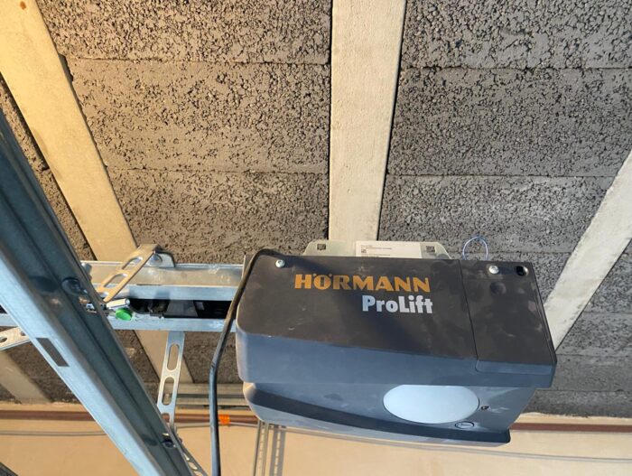 WhatsApp Image 2024-07-28 at 18.09.14 Na stronie internetowej znajduje się zdjęcie przedstawiające otwieracz drzwi garażowych Hörmann ProLift bezpiecznie zamontowany na suficie. Otwieracz jest przymocowany do solidnej metalowej szyny, bezproblemowo zintegrowanej z betonową i drewnianą konstrukcją belek sufitu, co jest przykładem profesjonalnej instalacji i harmonii konstrukcyjnej.