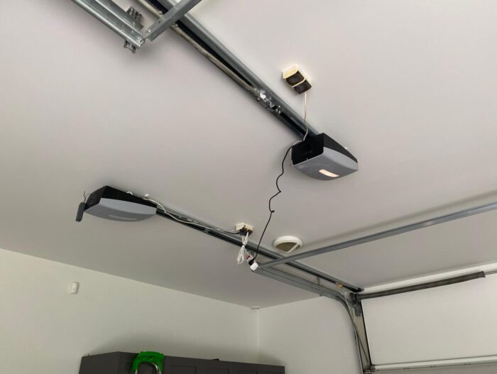 WhatsApp Image 2024-07-28 at 18.11.02 Na zdjęciu widać garaż wyposażony w montowany na suficie otwieracz drzwi, z widocznym okablowaniem. Do solidnych metalowych szyn przymocowane są dwa silniki, które ułatwiają otwieranie i zamykanie drzwi. W pobliżu znajduje się czujnik dymu dla większego bezpieczeństwa, a w tle widać szafkę jako część zestawu do przechowywania. Ta ilustracja nadaje się do witryny internetowej poświęconej usługom naprawy bram, rolet i barier w Warszawie.