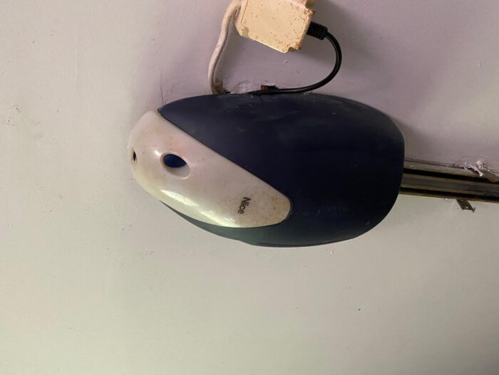 WhatsApp Image 2024-07-28 at 18.22.33 Na zdjęciu widać zbliżenie zabytkowego czarno-białego urządzenia sterującego bramą zamontowanego na ścianie, z odsłoniętym okablowaniem. Powierzchnia urządzenia wydaje się lekko zużyta, co dodaje mu charakteru. Wtyczka jest podłączona w prawym górnym rogu, a na urządzeniu wyraźnie widać słowo „Nice”.