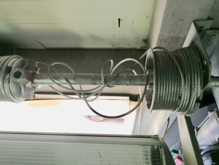 diploma (1) Na zdjęciu widać szczegółowy widok sprężyny skrętnej i systemu linek metalowej bramy garażowej. Mocno zwinięte linki są nawinięte wokół szpul połączonych ze sprężynami, które są zamocowane na metalowym pręcie umieszczonym nad bramą. Niektóre z tych linek wydają się być luźne lub splątane, co wskazuje na potencjalne problemy, które mogą wymagać naprawy.