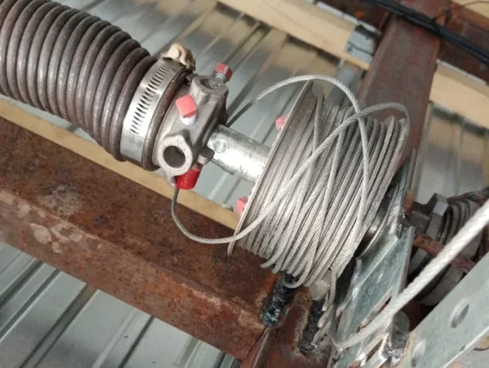 diploma (2) Obraz na stronie internetowej przedstawia szczegółowy widok metalowego kabla zwiniętego wokół bębna. Ta konfiguracja jest połączona z falistym wężem metalowym zabezpieczonym zaciskami. Urządzenie jest zainstalowane na zwietrzałej, zardzewiałej metalowej ramie, ustawionej na tle falistego metalu, co sugeruje jego rolę w środowisku przemysłowym lub mechanicznym.