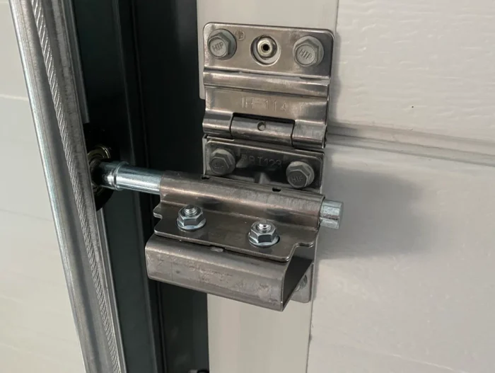 diploma Obraz na stronie internetowej przedstawia szczegółowy widok metalowego mechanizmu zawiasowego na białych drzwiach garażowych. Można zobaczyć śruby i cylindryczny pręt, które zabezpieczają drzwi, a także widoczne mocowania i metalowe elementy, odzwierciedlające kunszt ekspercki niezbędny do naprawy bram, rolet i barier w Warszawie.