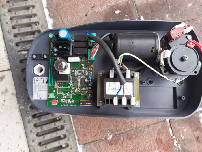 diploma (3) Obraz przedstawia skomplikowane wnętrze otwartej bramy elektronicznej lub urządzenia barierowego, podkreślając jego złożoną budowę obwodów. Widok ujawnia płytkę drukowaną ozdobioną szeregiem komponentów, w tym połączonymi przewodami, silnikiem i transformatorem. Całe urządzenie jest umieszczone na teksturowanej powierzchni, obok której znajduje się metalowa krata, odzwierciedlająca wiedzę techniczną oferowaną na tej warszawskiej stronie internetowej poświęconej naprawie bram i rolet.