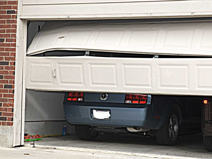 garage door repair-1000 Obraz na stronie internetowej przedstawia uszkodzoną bramę garażową, która jest wygięta i częściowo otwarta, gdy próbuje zamknąć się nad zaparkowanym w środku samochodem. Widoczny jest tył pojazdu, a po lewej stronie sceny znajduje się czerwony mur z cegły. Ta wizualizacja podkreśla potrzebę usług naprawy bram w Warszawie.