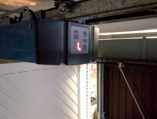 hq720 Obraz na naszej stronie internetowej przedstawia otwieracz drzwi garażowych zamontowany na suficie, wyposażony w wyświetlacz cyfrowy wskazujący literę „L”. Tło ukazuje część drzwi garażowych, podkreślając nasze doświadczenie w naprawach bram, rolet i barier w Warszawie.