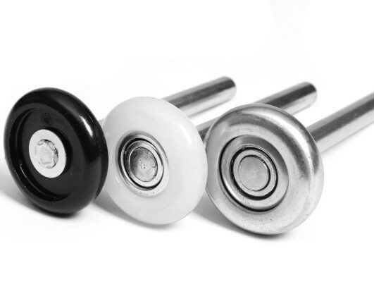 product-hardware-rollers Na stronie internetowej poświęconej naprawie bram, rolet i barier w Warszawie, na obrazie przedstawiono trzy rolki drzwi prysznicowych ustawione poziomo. Pierwsza rolka po lewej jest czarna, po niej biała w środku, a na końcu metaliczna rolka po prawej. Każda rolka ma metalową oś i jest umieszczona na prostym białym tle.