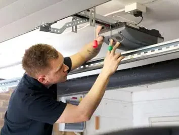 remont-sec-vorot-statya-avtoproezd-11 Na stronie internetowej poświęconej naprawom bram, rolet i barier w Warszawie, na zdjęciu widać osobę w czarnej koszuli, która pilnie naprawia lub instaluje mechanizm otwierania drzwi garażowych. Trzymając śrubokręt w ręku, osoba ta uważnie patrzy na urządzenie, pracując w garażu.