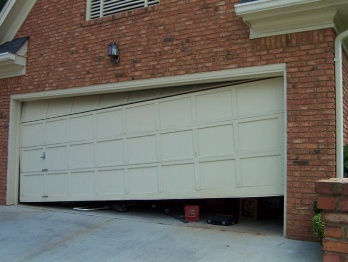 rg6 Obraz na stronie internetowej przedstawia ceglany dom z białymi drzwiami garażowymi, które są częściowo otwarte, wygięte i niewspółosiowe z torem. Wewnątrz garażu widoczne są różne przedmioty, w tym skrzynka narzędziowa i opona rowerowa.