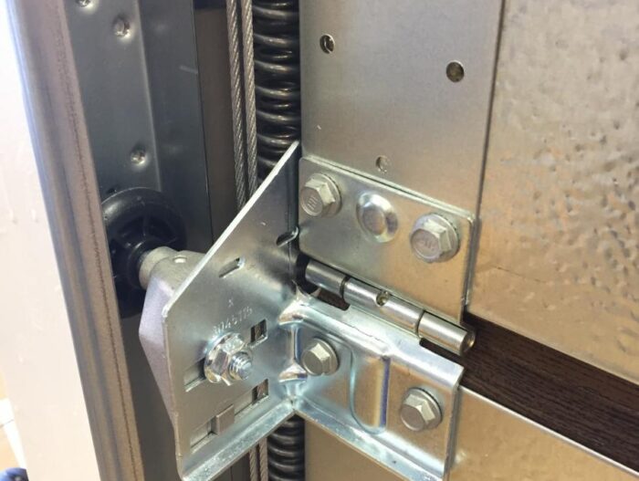 roliki_v_napravlyayushchey Na zdjęciu widać szczegółowy widok metalowego zawiasu drzwi garażowych ze zintegrowanym mechanizmem sprężynowym i przymocowanym kablem. Zawias jest mocno przymocowany śrubami do panelu, łącząc się bezproblemowo z drzwiami garażowymi. Ta ilustracja podkreśla kluczowe elementy mechaniczne, które są integralną częścią funkcjonalnego systemu drzwi garażowych, idealne dla osób zainteresowanych naprawą bram, rolet lub barier w Warszawie.