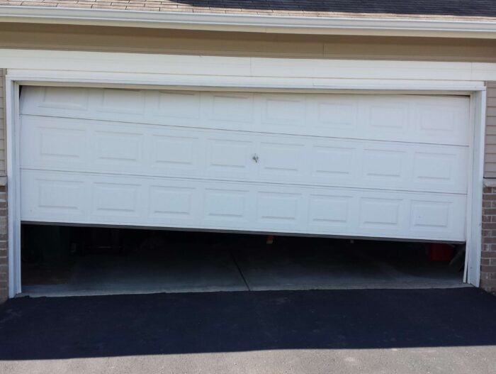 sl1 Na zdjęciu widać drzwi garażowe wymagające naprawy, ustawione pod nierównym kątem, z górnym lewym rogiem uniesionym nad prawym górnym. Ten garaż jest częścią beżowego domu akcentowanego cegłami i przylega do świeżo wybrukowanego czarnego asfaltu podjazdu.