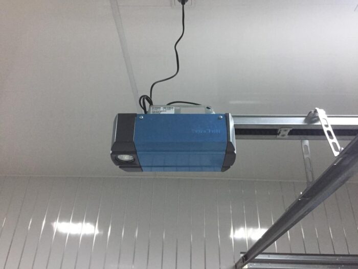 supramatic3 Obraz na stronie internetowej przedstawia niebiesko-szary otwieracz drzwi garażowych bezpiecznie zamontowany na suficie. Widoczny jest czarny przewód zasilający wychodzący z otwieracza, zapewniający zasilanie do jego działania. Okoliczny sufit i ściany są pomalowane na czysty, gładki biały kolor, odzwierciedlający schludne i profesjonalne otoczenie typowe dla usług naprawy bram, rolet i barier w Warszawie.