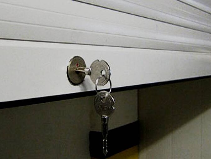 vidy-zamkov-dlya-rolstaven Na zdjęciu widać częściowo otwarte metalowe drzwi garażowe z poziomymi listwami, które podkreślają ich solidną konstrukcję. Zestaw kluczy zwisa z zamka na panelu drzwi, gotowy do użycia. Słabe oświetlenie tworzy stonowaną atmosferę, a część sąsiedniej ściany jest widoczna, co dodaje kontekstu do sceny uchwyconej na tej warszawskiej stronie internetowej poświęconej naprawie bram, rolet i barier.