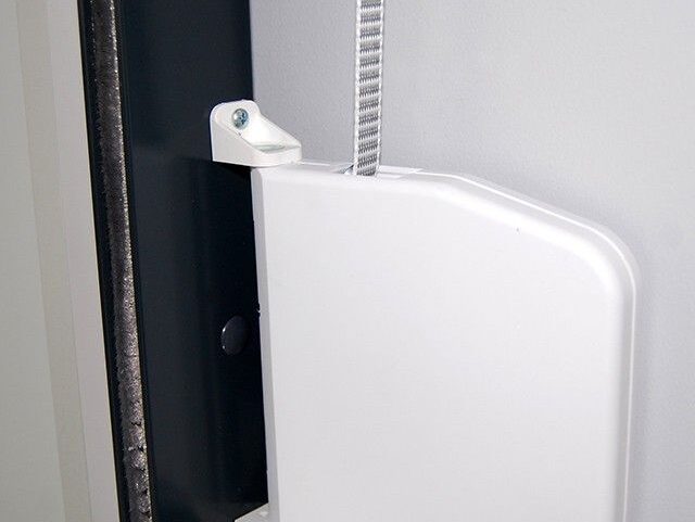 zwijacz-uniwersalny-do-rolet-zewnetrznych Obraz na naszej stronie internetowej, specjalizującej się w naprawie bram, rolet i barier w Warszawie, przedstawia szczegółowy widok białej plastikowej osłony zabezpieczonej solidnym metalowym paskiem. Ten element jest mocno zamontowany na ciemnej metalowej ramie, na tle szarej ściany. Wygląda jak integralna część mechanicznego systemu lub urządzenia.