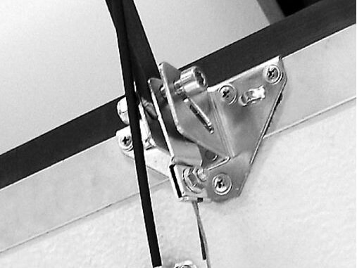 125_2 Czarno-biały obraz przedstawia szczegółowy widok metalowego zawiasu bezpiecznie przymocowanego do narożnika za pomocą śrub, wraz z przymocowanym kablem lub drutem. Elementy spoczywają na płaskiej powierzchni, ukazując skomplikowane detale typowe dla napraw bram, rolet i barier w Warszawie.