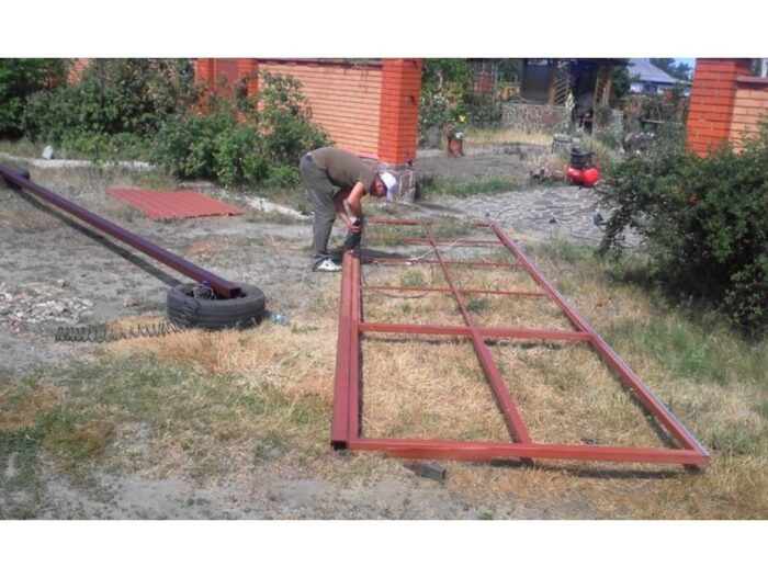 2015-06-20-11.03.57 Osoba jest widoczna podczas montażu lub naprawy sporej metalowej ramy bramy na zewnątrz, która jest umieszczona na ziemi w pobliżu ceglanej konstrukcji. Wokół znajduje się wiele różnych narzędzi i materiałów. Trawa w okolicy wydaje się sucha i nierówna, co wskazuje na potrzebę konserwacji.