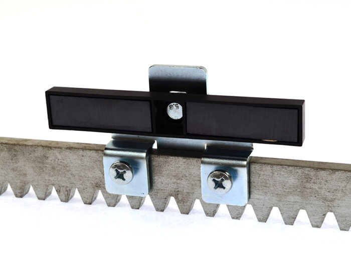 DSC_01131 Obraz na stronie internetowej przedstawia czarny prostokątny czujnik bezpiecznie przymocowany do metalowego pręta za pomocą dwóch śrub. Jest on umieszczony obok krawędzi przypominającej zęby piły, z wyraźnym białym tłem podkreślającym szczegóły komponentów.