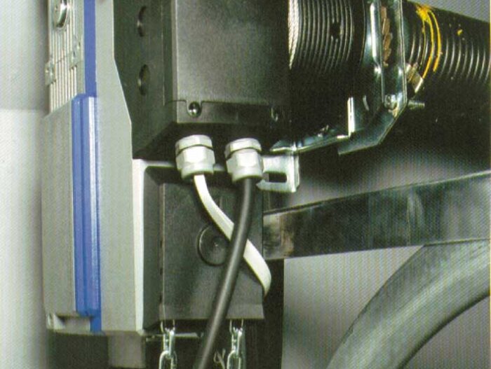 Industrietorantrieb-3 Obraz na stronie internetowej poświęconej naprawom bram, rolet i barier z siedzibą w Warszawie przedstawia szczegółowy widok komponentu maszyny przemysłowej. Zawiera czarną skrzynkę połączoną wieloma kablami, a także solidny metalowy łańcuch i starannie zwinięty wąż. Ten zespół jest bezpiecznie przymocowany do metalowej ramy, co sugeruje jego rolę jako integralnego elementu większego systemu mechanicznego lub sprzętu.