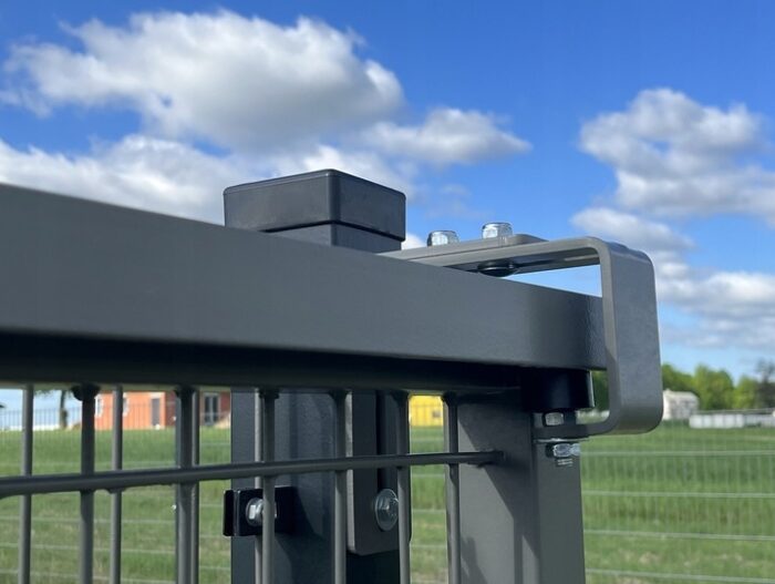 Uchwyt-katownik-rolki-prowadzacej-bramy-przesuwnej-ocynk Szczegółowy widok metalowej konstrukcji bramy wyposażonej w śrubę i zacisk, umieszczonej na tle bujnego zielonego pola i błękitnego nieba usianego rozproszonymi chmurami, zaprezentowany na stronie internetowej firmy specjalizującej się w naprawie bram, rolet i barier w Warszawie.