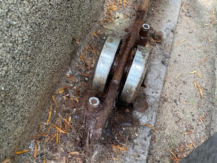 WhatsApp Image 2024-08-04 at 14.19.16 Szczegółowy widok skorodowanego metalowego mechanizmu rolkowego na tle kamiennej ściany, umieszczonego pośród rozrzuconych igieł sosnowych i ziaren piasku. Obraz ten podkreśla potrzebę profesjonalnych usług konserwacji i naprawy bram, rolet i barier w Warszawie.