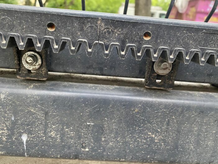 WhatsApp Image 2024-08-04 at 14.50.00 Obraz prezentowany na naszej stronie internetowej poświęconej naprawie bram, rolet i barier w Warszawie uwydatnia szczegółowy widok zakurzonego czarnego elementu mechanicznego z zazębiającymi się ząbkami, mocno przytrzymywanymi na miejscu przez solidne metalowe śruby. Tło jest delikatnie nieostre, odsłaniając ślady otaczającej zieleni obok różowych i szarych elementów.