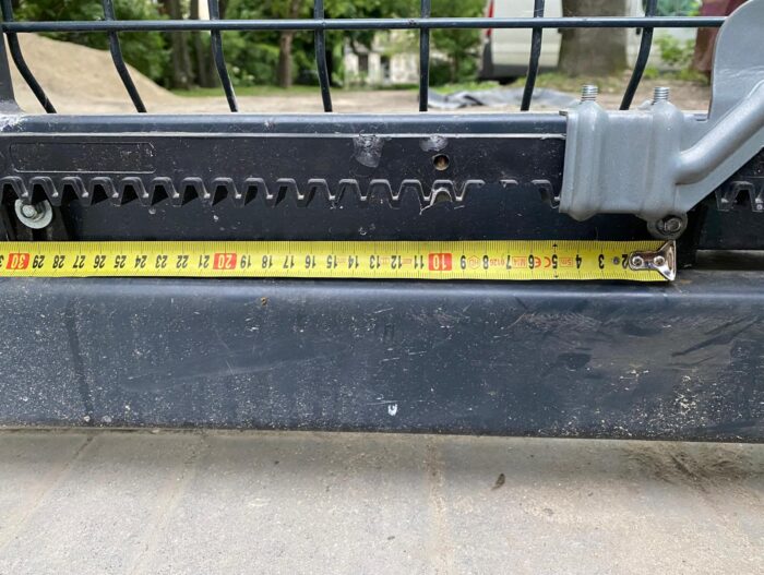 WhatsApp Image 2024-08-04 at 14.50.21 Obraz na stronie internetowej usług naprawy bram, rolet i barier w Warszawie przedstawia szczegółowe zbliżenie metalowego komponentu z kołami zębatymi i śrubami. Podświetlona na żółto taśma miernicza jest rozciągnięta na komponencie, wyraźnie pokazując pomiary w centymetrach. W tle widać siatkę drucianą na tle otoczenia zewnętrznego.