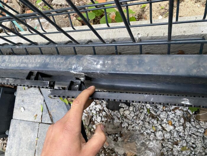 WhatsApp Image 2024-08-04 at 14.50.38 Na zdjęciu widać rękę wskazującą na uszkodzony kawałek plastiku na metalowym ogrodzeniu. W scenerii znajdują się żwir, rozrzucone liście i kostka brukowa. Zdjęcie jest prezentowane na stronie internetowej, która specjalizuje się w naprawie bram, rolet i barier w Warszawie.