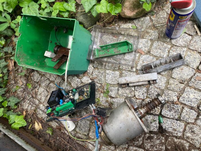 WhatsApp Image 2024-08-04 at 14.51.30 Obraz przedstawia scenę z witryny internetowej zajmującej się naprawą bram, rolet i barier z siedzibą w Warszawie. Na brukowanej powierzchni otoczonej zielenią rozrzucone są różne elementy elektroniczne i metalowe części. Najbardziej widoczne są zielone metalowe pudełko, kilka przewodów, płytki drukowane i puszka smaru.