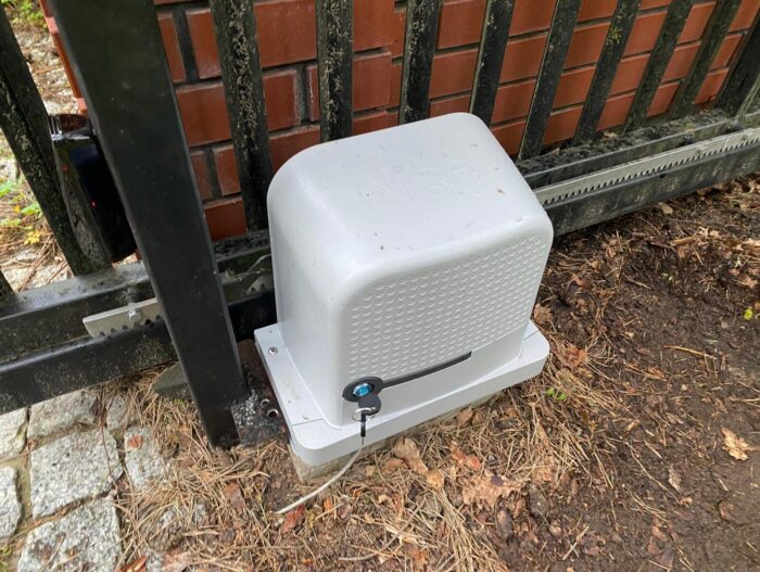 WhatsApp Image 2024-08-04 at 15.19.58 Na zdjęciu widać biały, prostokątny silnik automatycznej bramy umieszczony na ziemi obok czarnej metalowej bramy i przylegający do ceglanego muru. Silnik zawiera dziurkę od klucza i wytłoczone logo na swojej powierzchni. Okoliczny obszar jest pokryty brudem i rozrzuconymi liśćmi, co dodaje naturalnego otoczenia. Ta instalacja jest przykładem fachowego rzemiosła w zakresie usług naprawy bram, rolet i barier oferowanych w Warszawie.