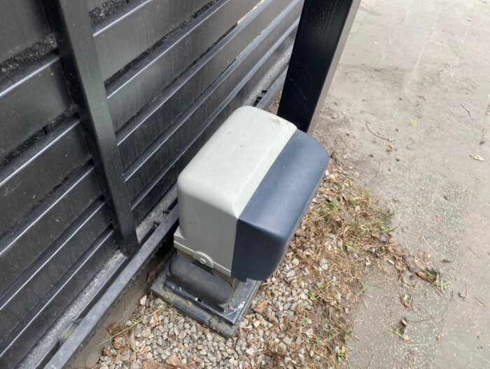 WhatsApp Image 2024-08-04 at 15.20.57 Obraz na stronie internetowej przedstawia elegancki czarno-szary otwieracz bramy z napędem umieszczony na żwirowym fundamencie obok solidnej metalowej bramy. Obszar wokół instalacji jest usiany małymi kamieniami i suszonymi liśćmi, co dodaje otoczeniu naturalnego akcentu.