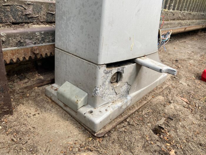 WhatsApp Image 2024-08-04 at 15.22.05 Obraz na stronie internetowej z siedzibą w Warszawie poświęconej naprawom bram, rolet i barier przedstawia zwietrzałą szarą metalową skrzynkę, umieszczoną na betonowej powierzchni obok ogrodzenia. Skrzynka wykazuje ślady rdzy i zużycia, z odsłoniętym metalowym prętem i postrzępionymi przewodami u podstawy, wszystko umieszczone w zakurzonym środowisku zewnętrznym.