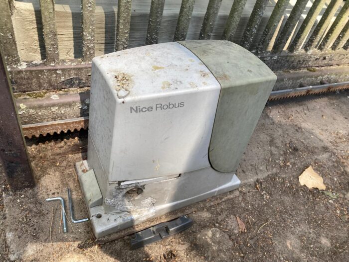 WhatsApp Image 2024-08-04 at 15.22.19 Na stronie internetowej poświęconej usługom naprawy bram, rolet i barier w Warszawie, obraz przedstawia zwietrzały szary silnik bramy Nice Robus zamontowany na betonowej podstawie. Silnik nosi ślady rdzy i jest umieszczony przed ogrodzeniem, a za nim widoczna jest metalowa szyna.