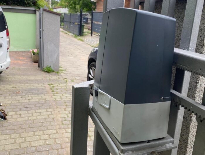 WhatsApp Image 2024-08-04 at 15.23.00 Na zdjęciu widać szczegółowy widok eleganckiego szarego i czarnego silnika bramy przesuwnej, bezpiecznie przymocowanego do solidnej metalowej ramy, umieszczonego obok brukowanego podjazdu. W tle widać biały samochód zaparkowany w pobliżu kilku doniczek z kwiatami, a ulica za nim jest ograniczona różnymi płotami, co przyczynia się do zadbanego wyglądu charakterystycznego dla warszawskich dzielnic.