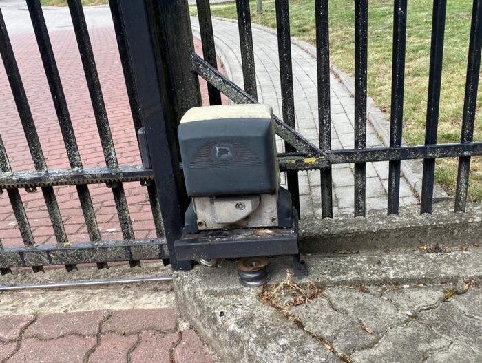 WhatsApp Image 2024-08-04 at 15.23.41 Na zdjęciu widać elegancką czarną metalową bramę z automatycznym mechanizmem przesuwnym, bezproblemowo zainstalowaną na betonowej nawierzchni. Otwieracz bramy jest umieszczony blisko ziemi, a widoczne są fragmenty ścieżki i przyległe obszary trawiaste. Ta konfiguracja przedstawia profesjonalne usługi naprawcze i instalacyjne dostępne w Warszawie dla bram, rolet i barier.