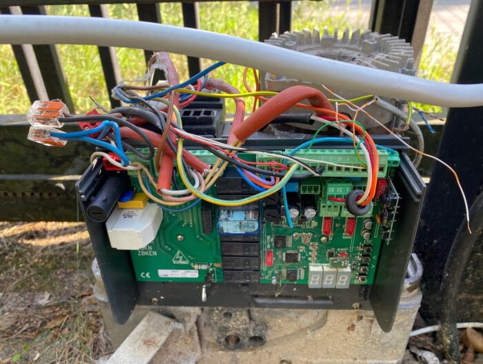 WhatsApp Image 2024-08-04 at 15.34.58 Obraz na naszej stronie internetowej przedstawia szczegółowy widok otwartej skrzynki sterowniczej wypełnionej szeregiem kolorowych przewodów misternie połączonych z płytką drukowaną. Ta skrzynka sterownicza jest bezpiecznie zamontowana na zewnątrz na solidnej metalowej bramie lub ogrodzeniu, z bujną trawą widoczną w tle, ucieleśniając istotę profesjonalnych usług naprawczych i konserwacyjnych bram, rolet i barier w Warszawie.
