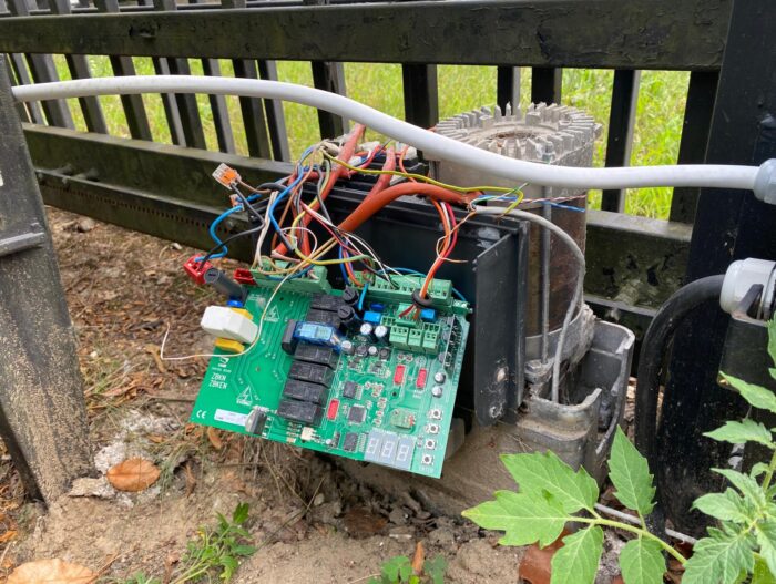 WhatsApp Image 2024-08-04 at 15.35.15 Obraz na stronie internetowej poświęconej naprawie bram, rolet i barier w Warszawie przedstawia zewnętrzną metalową konstrukcję z odsłoniętą płytką drukowaną. Ta płytka drukowana jest usiana różnymi elementami elektronicznymi i skomplikowanymi splotami kolorowych przewodów. Całość umieszczono na tle brudu i roślinności, podkreślając środowisko pracy, w którym często przeprowadzane są te naprawy.