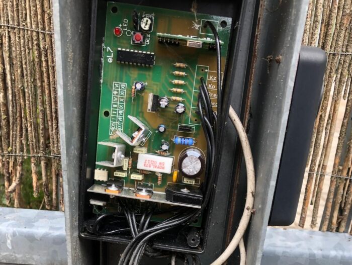 WhatsApp Image 2024-08-04 at 15.36.00 Szczegółowy obraz pokazuje otwartą skrzynkę sterowniczą, odsłaniając płytkę drukowaną gęsto wypełnioną elementami, takimi jak przewody, kondensatory i rezystory. Ta skomplikowana konstrukcja jest bezpiecznie zamontowana na solidnej metalowej ramie, ustawionej na tle drewnianego ogrodzenia. Zdjęcie doskonale ilustruje drobiazgową pracę związaną z naprawą bram, rolet i barier w Warszawie.