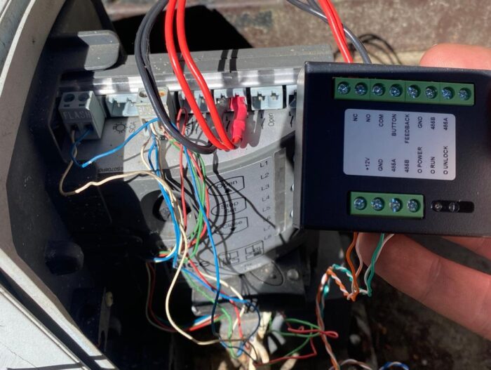 WhatsApp Image 2024-08-04 at 15.38.53 Obraz na naszej stronie internetowej poświęconej usługom naprawy bram, rolet i barier w Warszawie przedstawia szczegółowe ujęcie skomplikowanej instalacji okablowania. Liczne kolorowe przewody są misternie podłączone do różnych zacisków na płytce drukowanej. Widać rękę trzymającą kompaktowe czarne urządzenie wyposażone w zielone bloki zacisków i wyraźnie oznaczone porty, takie jak „OUTLOCK” i „INHIBIT”.